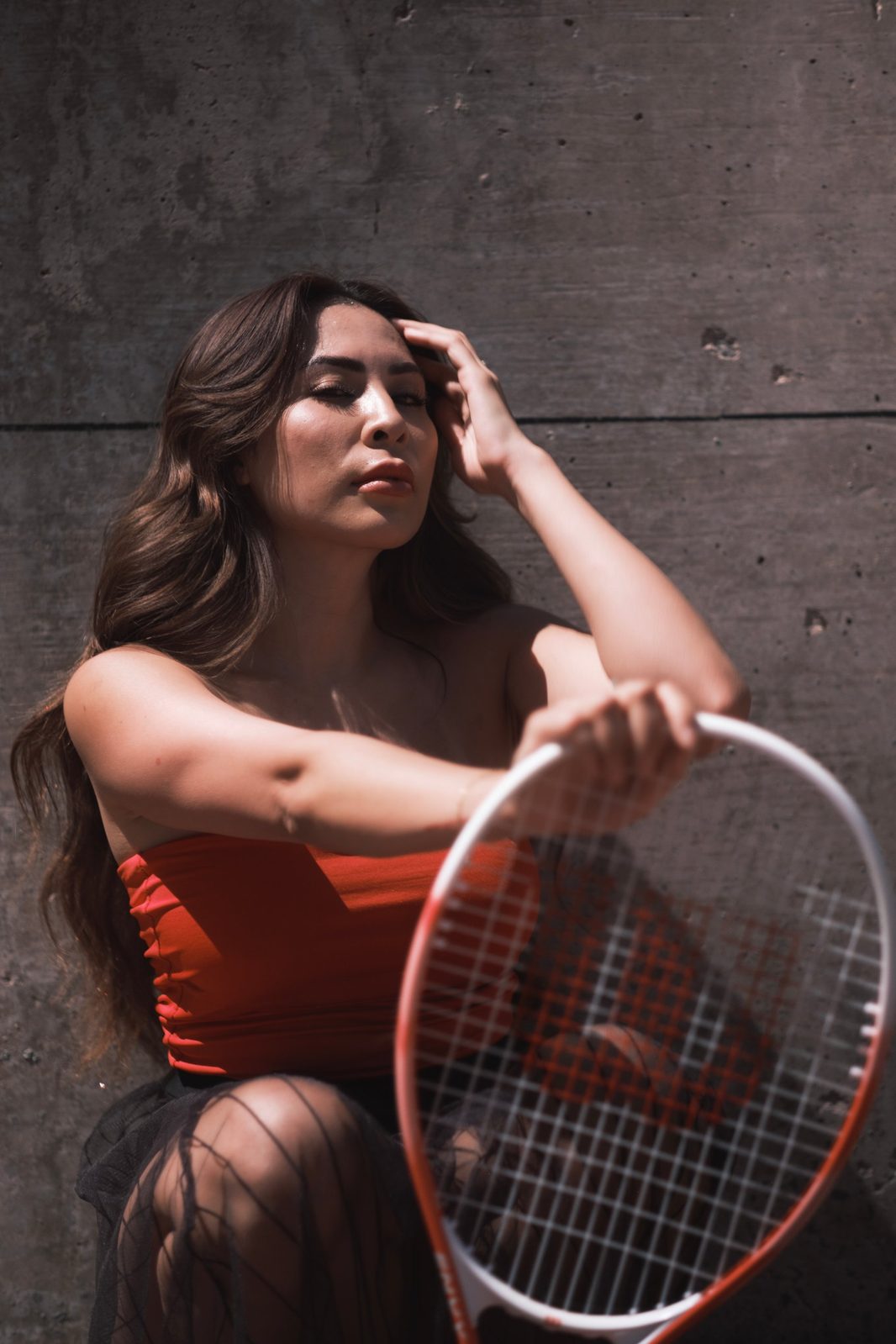 Portrait seated against a concrete wall in a red top, holding a tennis racket
